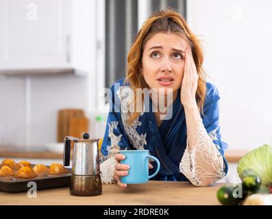 Junge Frau, die an Migräne leidet, Kaffee in der Küche trinkt Stockfoto