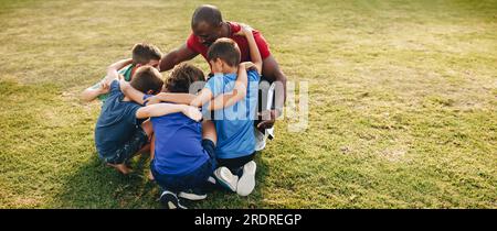 Schulkinder drängen sich mit ihrem Trainer auf einem Sportplatz. Der Trainer spricht ein motivierendes Team mit einer Gruppe von Grundschulkindern. Sport e Stockfoto