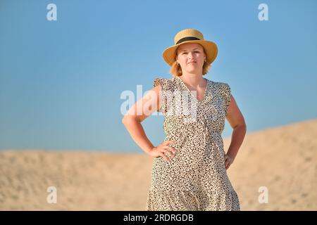 Junge Frau in langem Kleid und Strohhut auf der Sanddüne. Glückliche europäische Frau Genießen Sie einen sonnigen Tag im Urlaub in der Wüste. Stockfoto