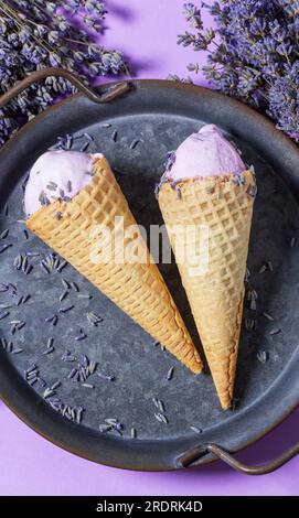 Zwei Lavendeleis in Waffle Cones auf einem Tablett und Blumensträuße aus trockenem Lavendel auf lila Hintergrund. Stockfoto