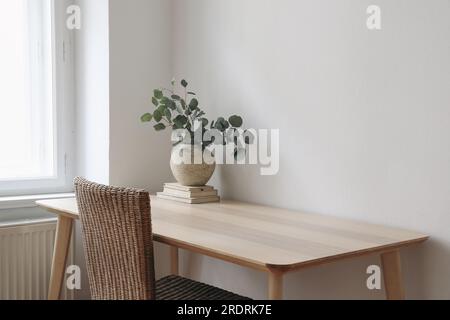 Stilvolle Innenausstattung. Wohnzimmer mit Vase, Eukalyptusäste, alte Bücher auf Holztisch. Rattanstuhl in der Nähe des Fensters. Minimalistisches Heimkonzept Stockfoto