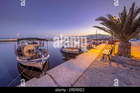 Fischerhafen-Szene in Griechenland mit Booten, Palmen und Motorrollern bei Nacht an einem wunderschönen ruhigen Sommertag Stockfoto