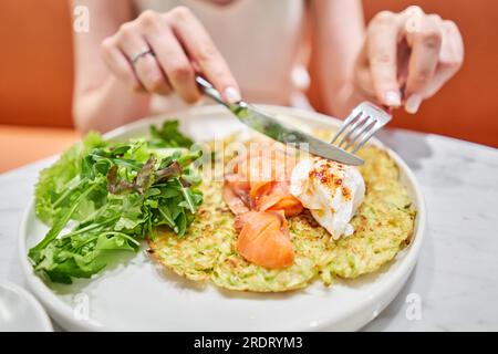 Frau schneidet Zucchini-Pfannkuchen mit Avocado, Käsesahne, Lachs und Ei. Gesundes Frühstück, Eiweiß. Restaurant-Gericht. Frühstück im Café bei Sonnenschein Stockfoto