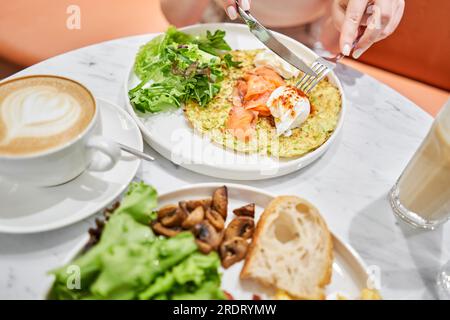 Frau schneidet Zucchini-Pfannkuchen mit Avocado, Käsesahne, Lachs und Ei. Gesundes Frühstück, Eiweiß. Restaurant-Gericht. Frühstück im Café bei Sonnenschein Stockfoto