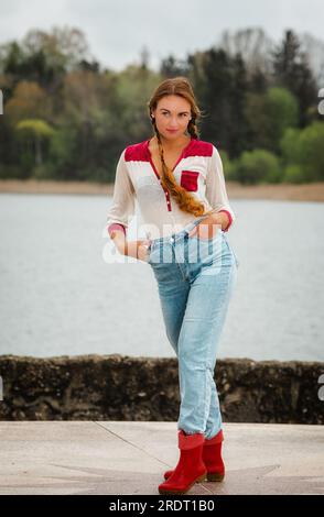 Junge Frau mit langen Haaren am Seeufer in Jeans und Gummistiefeln. Stockfoto