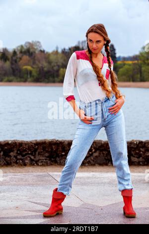 Junge Frau mit langen Haaren am Seeufer in Jeans und Gummistiefeln in Modellstellung Stockfoto