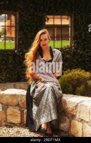 Schöne Frau in festlichem Dirndl, die an einer Steinwand sitzt Stockfoto