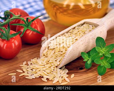 Nicht gekochte griechische Nudeln, Kritharaki in Holzlöffel auf Schneidebrett für Olivenholz mit frischen Tomaten und Oregano Stockfoto