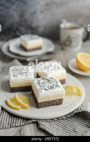 Cremige Zitronen- und Mohnriegel auf einem weißen Teller, dekoriert mit frischen Zitronenscheiben. Grauer Hintergrund und abgezogene Serviette. Eine Tasse Kaffee. Stockfoto