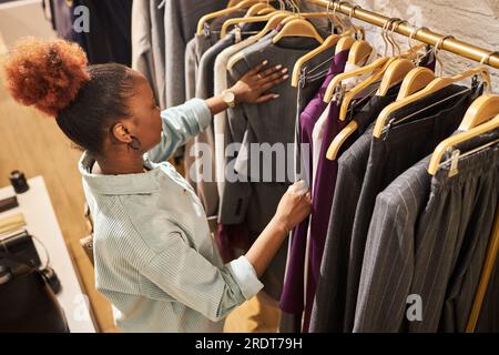 Hochwinkel-Porträt einer jungen afroamerikanischen Frau, die Kleidung auf einem Kleiderständer im Kleiderladen durchsucht, Kopierraum Stockfoto