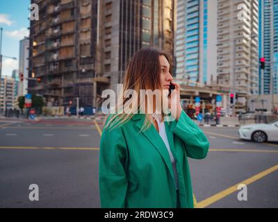Junge Frau im grünen Anzug geht durch die Straßen von Dubai, Dubai Marina District. Reisekonzept der Vereinigten Arabischen Emirate. Die Vorstellung von erfolgreichem Auswanderer, Umzug in ein anderes Land, Arbeitsvisum Stockfoto
