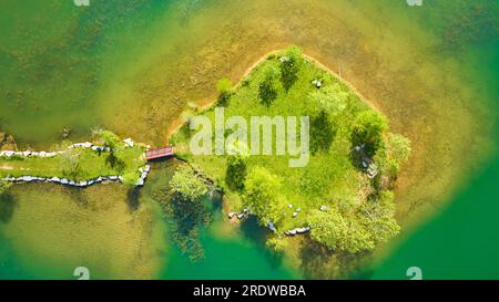 Aus der Luft über den türkisgrünen Teich mit kleiner Insel und Landbrücke, die zur roten Wanderbrücke führt Stockfoto