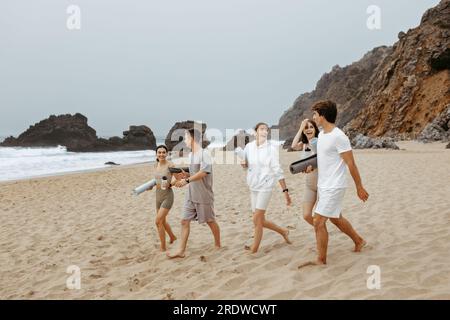 Eine Gruppe glücklicher junger Leute, die mit Yogamatten am Strand spazieren gehen, sich auf ein frisches Training an der Küste vorbereiten, freier Platz Stockfoto