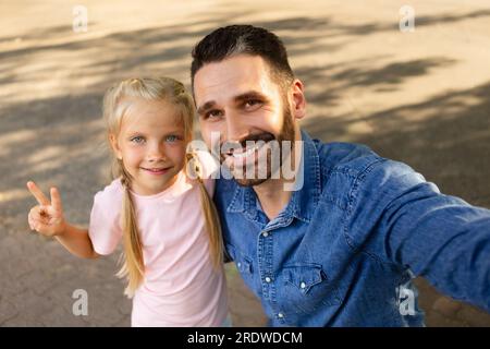 Ein glücklicher weißer Vater mittleren Alters, der Selfie mit seiner süßen Tochter macht, ein Mädchen, das eine friedliche Geste zeigt, im Park spaziert Stockfoto