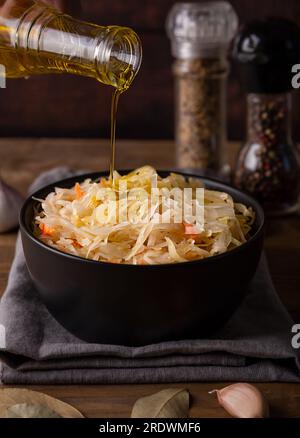 Dunkle Schüssel mit saurem Sauerkraut auf dem Schneidebrett auf holzbraunem Hintergrund – Nahaufnahme. Daneben gibt es Kohl, Karotten, Salz und Öl Stockfoto