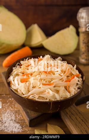 Dunkle Schüssel mit saurem Sauerkraut auf dem Schneidebrett auf holzbraunem Hintergrund – Nahaufnahme. Daneben gibt es Kohl, Karotten, Salz Stockfoto