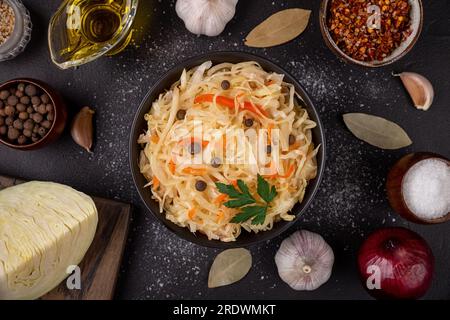 Schwarze Schüssel mit saurem Sauerkraut dekorierte Petersilie und Pfeffer auf dunklem Hintergrund liegend mit Zutaten wie frischem Kohl, Öl und Knoblauch Stockfoto