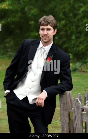 Der Bräutigam lehnt sich an seinem Hochzeitstag vor der Kirche an einen rustikalen Holzzaun. Er trägt einen schwarzen Smoking mit weißer Weste, Hemd und Krawatte. Er ist es Stockfoto