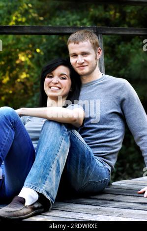 Junge Paare genießen einen ruhigen Abend am See. Beide lehnen sich gegen ein rustikales, hölzernes Dock nahe aneinander. Sie lächeln und sind glücklich. Stockfoto