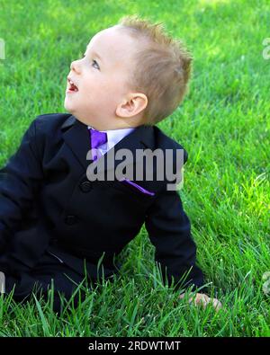 Der kleine Junge blickt in Erstaunen und Staunen auf. Sein Mund ist offen und er trägt einen schwarzen Anzug und eine lila Krawatte. Er liegt draußen auf dem Gras. Stockfoto