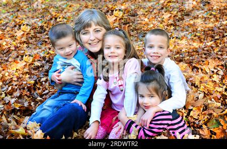 Als sie Herbstblätter harkte, hielt diese Großmutter an, um all ihre Enkel zu umarmen. Sie sitzen auf einem Laubhaufen und lächeln glücklich. Die Kinder einschl Stockfoto