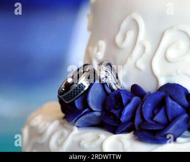 Zwei Eheringe schmiegen Amoungblaue Rosen auf einer Hochzeitstorte. Der Kuchen ist abgestuft und mit Wirbeln dekoriert. Stockfoto