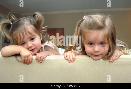 Zwei entzückende kleine Mädchen schauen über die Oberseite des Sofas in ihrem Haus. Schwestern, diese beiden sehen aus wie Ärger. Der jüngste hat Zöpfe. Stockfoto
