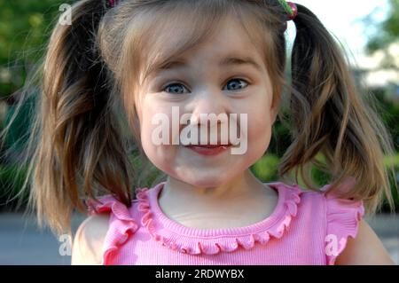 Das kleine Mädchen versucht, ihr Kichern zu halten, während sie ihr Gesicht in ein fröhliches Grinsen versetzt. Sie hat Zöpfe und ein rosa Hemd. Stockfoto