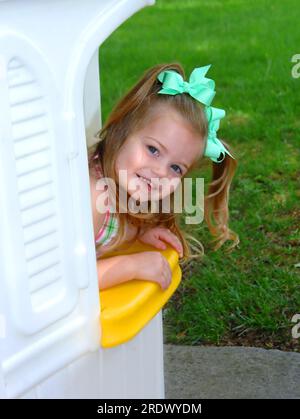 Das kleine Mädchen lehnt sich aus ihrem weißen Spielhaus und lädt alle ein, mit ihr zu spielen. Sie hat lange blonde Locken und grüne Bänder. Stockfoto