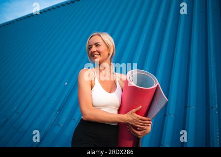 Porträt einer mittelalterlichen wunderschönen blonden Frau mit Sportmatte auf blauem Hintergrund. Gesunder, aktiver Lebensstil, Yoga-Workout Stockfoto