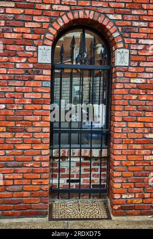 Rote Backsteinwand mit schwarzem Metalltor, der die Tür blockiert, jetzt mit Fenster und Bodenmatte aus Metall versteinert Stockfoto