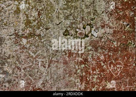 Graue Betonwand mit rotem Rost und grünem Moos mit zerbröckelnden Teilen und geschnitzten Herzen Stockfoto