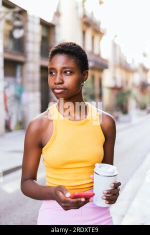 Rücksichtsvolle afrikanerin, die auf der Straße steht und wegschaut. Sie hält eine wiederverwendbare Kaffeetasse und ein Smartphone in der Hand. Stockfoto