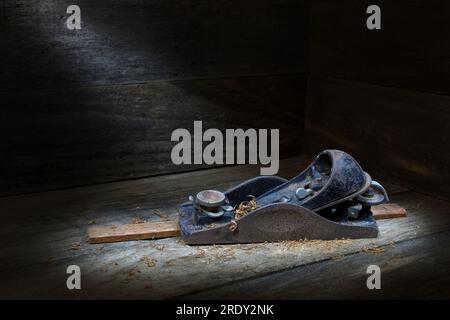 Ein kleines, altmodisches, rostiges, eisernes Tischlerblock-Handflugzeug mit Profil auf einem Holzstreifen mit Spänen, in sanfter, dunkler Stimmungsbeleuchtung mit Kopierbereich Stockfoto