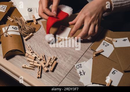 Eine nicht wiedererkennbare junge Frau klebt die Nummer auf die Handwerkstasche, wird mit einer Wäscheklammer befestigt. Weiblich macht Kraftpapier für hausgemachte Adventskalender mit eigenen Händen Schritt für Schritt selbst gemacht. Vorbereitung auf das weihnachtskonzept. Saisonale Aktivitäten für Kinder, Familienurlaub im Winter. Umweltfreundlich Geschenke. Öffnen Sie das Paket jeden Tag Stockfoto