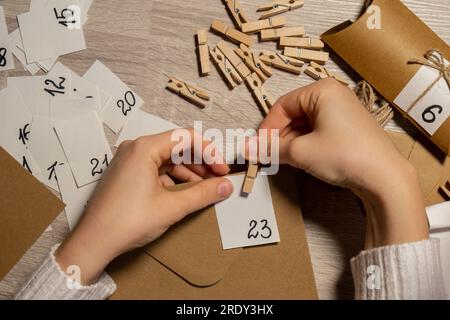 Eine nicht wiedererkennbare junge Frau klebt die Nummer auf die Handwerkstasche, wird mit einer Wäscheklammer befestigt. Weiblich macht Kraftpapier für hausgemachte Adventskalender mit eigenen Händen Schritt für Schritt selbst gemacht. Vorbereitung auf das weihnachtskonzept. Saisonale Aktivitäten für Kinder, Familienurlaub im Winter. Umweltfreundlich Geschenke. Öffnen Sie das Paket jeden Tag Stockfoto