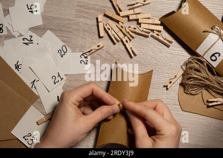 Eine nicht wiedererkennbare junge Frau klebt die Nummer auf die Handwerkstasche, wird mit einer Wäscheklammer befestigt. Weiblich macht Kraftpapier für hausgemachte Adventskalender mit eigenen Händen Schritt für Schritt selbst gemacht. Vorbereitung auf das weihnachtskonzept. Saisonale Aktivitäten für Kinder, Familienurlaub im Winter. Umweltfreundlich Geschenke. Öffnen Sie das Paket jeden Tag Stockfoto