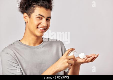 Fröhlicher junger, lächelnder Kerl, der sich eine Gesichtswäsche an die Finger legt und sich darauf vorbereitet, das Schönheitsprodukt auf sein Gesicht aufzutragen. Schwärziger Mann mit schwarzen Locken benutzt Stockfoto