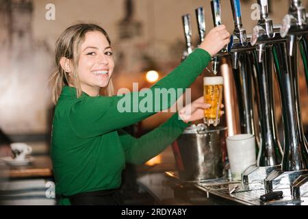 Fröhliche junge Kellnerin, die Biergläser vom Fass an der Bar füllte Stockfoto