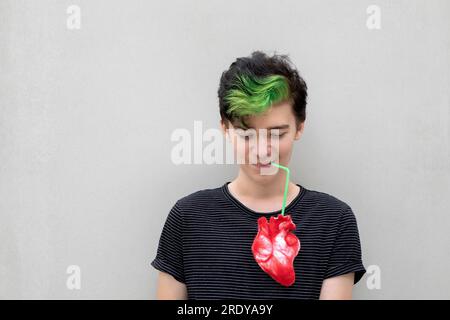 Ein Teenager mit grünem Haar trinkt aus dem Herzen vor grauem Hintergrund Stockfoto