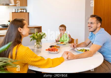 Ein Junge sieht lächelnde Eltern an, die sich an einem Esstisch Händchen halten Stockfoto