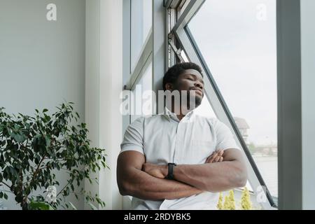 Junger Geschäftsmann mit geschlossenen Augen, der sich im Büro an ein Glasfenster lehnt Stockfoto