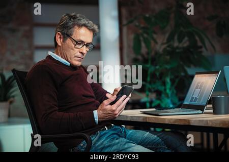 Architekt, der im Büro neben einem Laptop sitzt und ein Smartphone verwendet Stockfoto