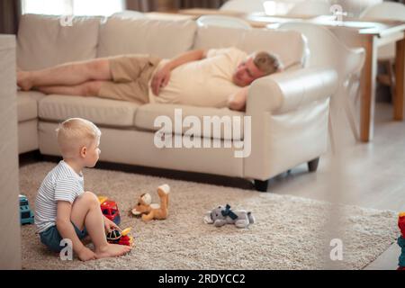 Der Sohn spielt mit Spielzeug und der Vater schläft zu Hause auf dem Sofa Stockfoto