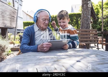 Seniorin trägt kabellose Kopfhörer, die zusammen mit dem Enkel einen Tablet-PC verwenden Stockfoto