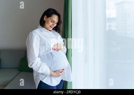 Lächelnde schwangere Frau, die zu Hause am Fenster stand Stockfoto