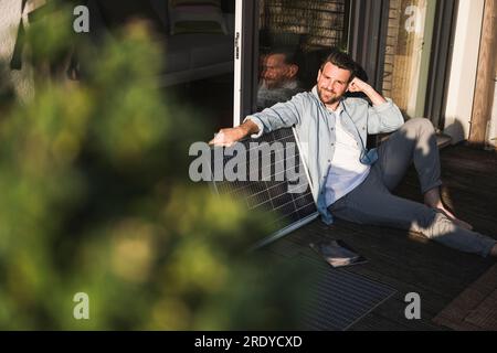 Lächelnder Geschäftsmann, der mit Solarpaneelen auf der Veranda sitzt Stockfoto