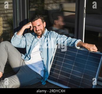 Glücklicher Geschäftsmann, der mit Solarpaneel auf der Veranda sitzt Stockfoto