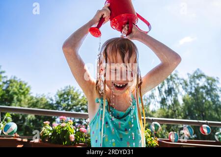 Lächelndes Mädchen, das Wasser aus der roten Gießkanne auf dem Balkon auf den Kopf gießt Stockfoto