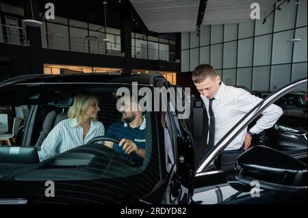 Der Verkäufer demonstriert den Käufern das Fahrzeuginnere. Ein weißes Paar sucht sich ein Auto aus. Stockfoto